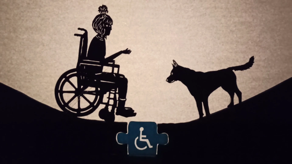Fotograma del cortometraje "Como pez en el agua". 
Títere de una niña en silla de ruedas cruzando de un lado a otro gracias a la accesibilidad, junto a la ayuda de su perro.